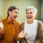 Elderly sisters watching comedy together indoors, laughing. Portrait of two beautiful Caucasian women relaxing at home, sitting on sofa, opening mouth wide opened, enjoying weekend, having fun
